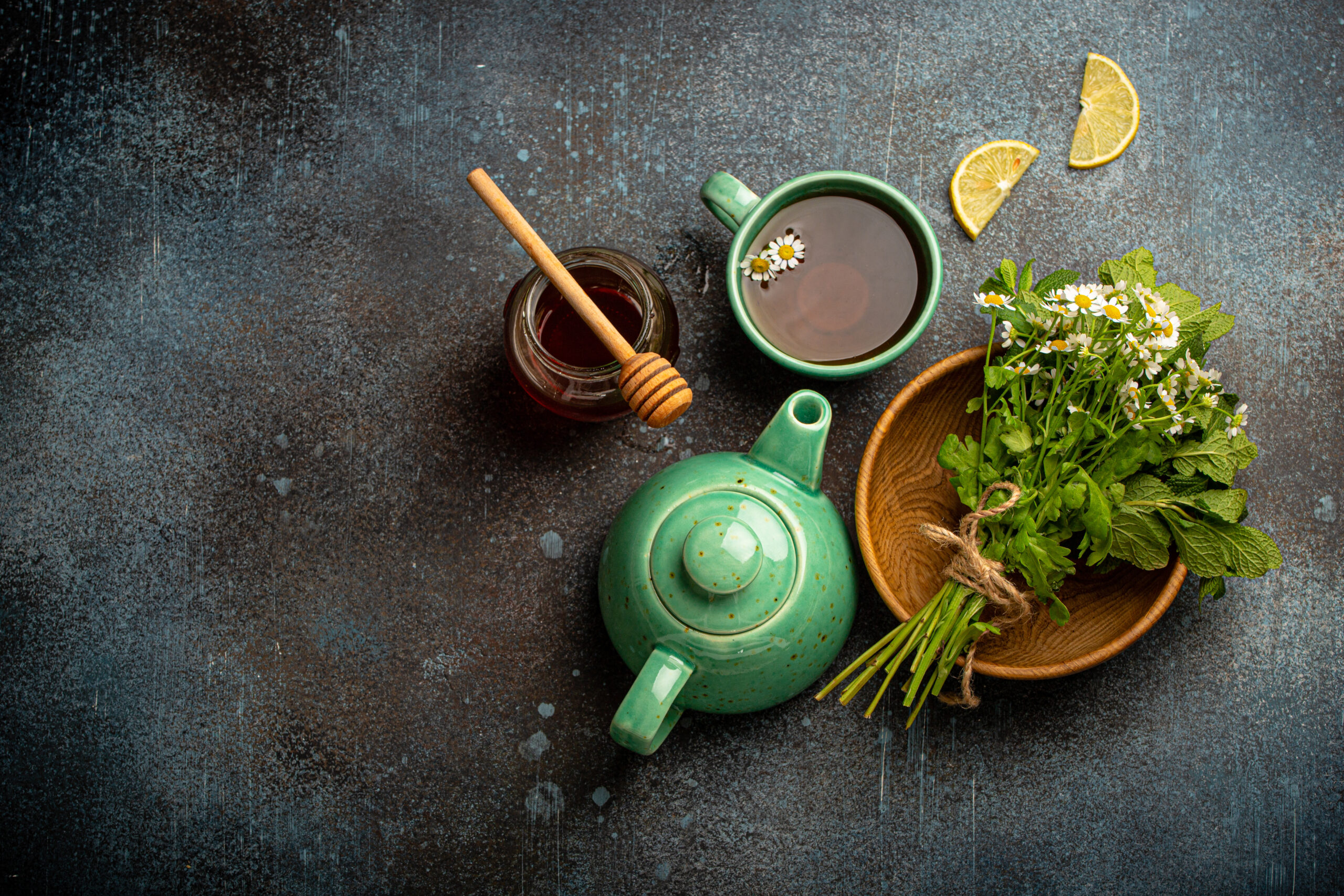 Herbal tea with camomile, fresh medical herbs and flowers, tea cup, tea pot, jar of honey, lemon top view on stone background, herbal alternative medicine and homeopathy remedy concept